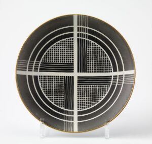  A modernist porcelain plate designed by Nora Gulbrandsen, featuring a dark background with a white grid circle at the center, bisected by perpendicular lines and highlighted with gold trim.