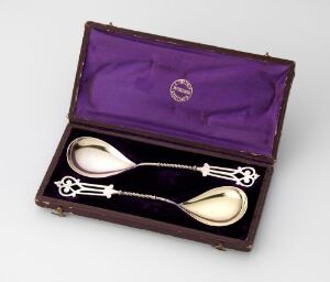  A set of two silver-gilded compote spoons designed by Oluf Tostrup, presented in an open dark purple-lined box. The spoons feature ornate handles with embossed designs and rest on fitted placeholders within the box, which showcases a circular emblem on the padded lid's lining.