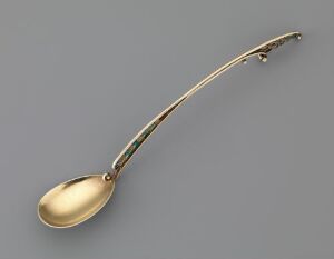  A gilded silver mocha spoon named "Mokkaskje" by artist J. Tostrup, with a long, curved handle and a golden hue, set against a neutral grey background.