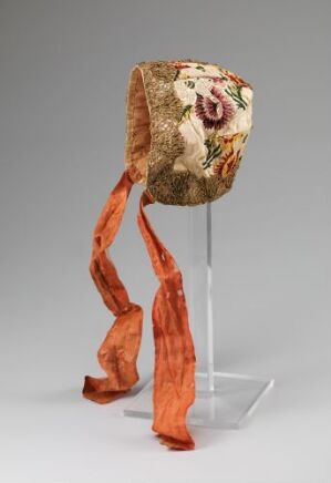  A sculptural art object with a cylindrical body showcasing vintage floral patterns in pink and green, and flowing ribbon-like burnt orange extensions, displayed against a neutral gray background. Artist name and title unknown.