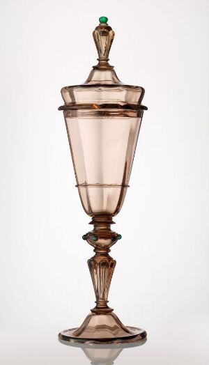  A smoky brown, glassware piece with a conical-shaped bowl and lid, featuring a slender stem with spherical elements, and a contrasting green round finial on top, against a light neutral background. Artist name and title are unknown.