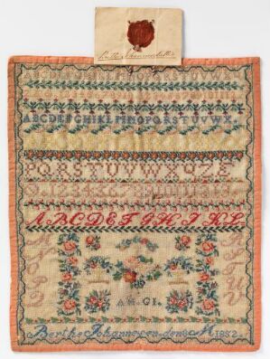  An antique needlework sampler with top-sealed paper, featuring multiple alphabets and a vibrant floral embroidery in red, blue, green, and pink on an off-white fabric, displaying needlework skills.