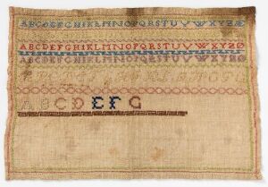  Antique beige linen sampler featuring multiple rows of alphabet letters in uppercase of varying sizes and styles, stitched in brown and yellow threads, a numerical row, a decorative motif, and a central, larger set of letters "C D E F G" in dark brown, with possible space for more