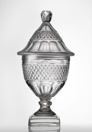  An intricately cut clear glass candy dish with a lid set against a light grey background. The dish has a faceted knob, diamond patterns on its dome-shaped lid, and a pedestal base, capturing light and displaying craftsmanship in glass