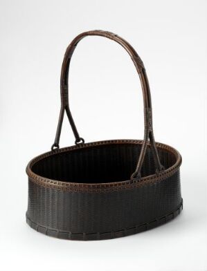  An oval-shaped dark brown woven basket with a single arched handle, exhibiting a ribbed texture, against a neutral gray background.