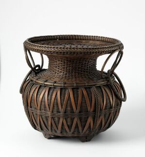 
 A beautifully woven round basket with a fluted opening, featuring intricate patterns created by the interplay of dark and light brown natural fibers, and two symmetrically-placed handles.