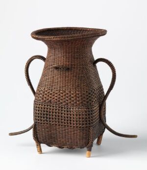  A woven brown basket with a flared open top, resembling a traditional pitcher, featuring symmetrical curved handles and standing on four small legs.