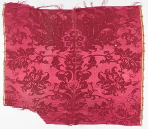 
 An antique fragment of deep red textile with a darker red symmetrical, ornate floral pattern, exhibiting signs of age and frayed edges, with a hint of yellow stitching on the top right corner.