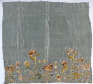  A fragment of an antique-looking textile with frayed edges, featuring an embroidered floral design in warm colors such as golden yellow, burnt orange, and soft green on a muted grey or pale blue background.