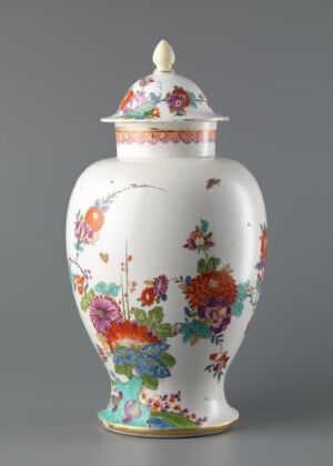  A white porcelain vase with a colorful floral design, featuring prominent flowers in shades of pink, orange, red, and purple amidst green foliage, topped with a rounded, pointed lid.