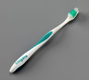  A toothbrush designed by Geir Øxseth with a white and teal handle featuring the Colgate logo, and bristles fading from white to teal to turquoise, against a gray background.