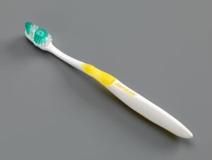  A "Sanodent" toothbrush designed by Geir Øxseth, featuring a white handle with a bright yellow accent and a green rubber grip at the base of the brush head, which has green bristles, all set against a light grey background.