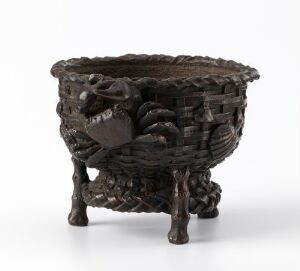  A dark-colored, small, intricately designed object resembling a woven basket or bowl with legs, featuring an animal head motif on one side and a texture that suggests traditional craftsmanship. The object appears to be made of a dark, metallic or ceramic material. Artistname and title are unknown.