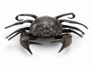  A sculpture combining a human skull with the body of a crab, featuring a metallic finish in shades of pewter and silver. The skull serves as the body with detailed features, and the crab's appendages are long and slender, with oversized pincers, all set against a white background.