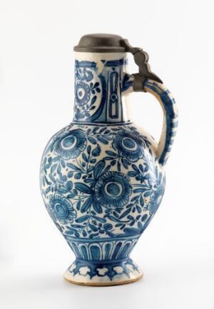  A blue and white ceramic jug with a metal lid, showcasing intricate floral patterns and ornamental designs, against a white background. The jug has a curved handle and stands on a flared base, with vibrant cobalt blue designs on a glossy white surface.