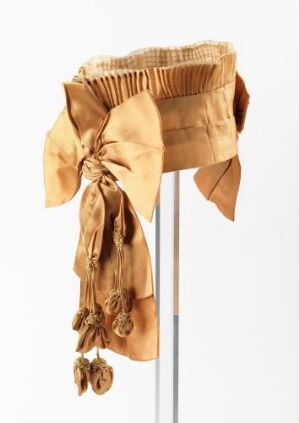  A vintage golden brown silk bonnet with detailed ruching and an oversized bow, featuring hanging rosette embellishments, displayed on a thin pole against a white background.