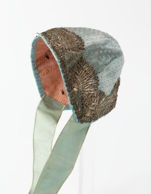  A traditional conical hat with metallic gold, silver, and bronze thread embellishments, decorated with muted blue and green fabric patches, with pale green ribbons extending from the back, all displayed against a white background.