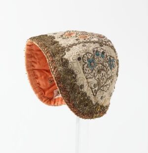  An intricately embroidered traditional cap with a floral and avian design in muted earth tones, accented with golds, blues, and greens, against a light gray background. The inside is lined with coral-colored fabric.