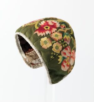  A traditional floral patterned bonnet in olive green with red, pink, yellow, and white flowers, lined with an off-white, fuzzy material, photographed against a white background. Artist name and title are unknown.