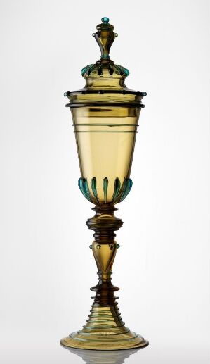  An intricate glass goblet with a spiraled yellow-green base and stem, turquoise curved prongs supporting a creamy yellow body, and a matching lid with turquoise accents against a light background.