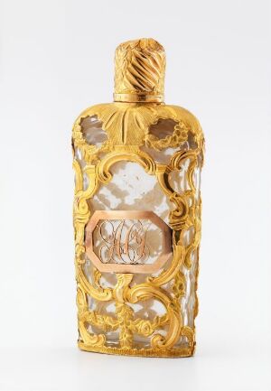  A gold-colored, ornate bottle with baroque-style decorations and a clear central panel featuring an ambiguous monogram or symbol, set against a soft white background.