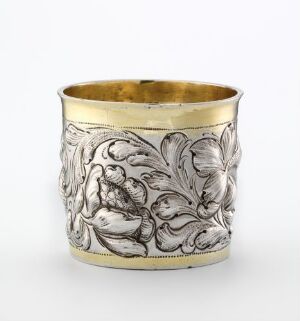  A cylindrical silver cup with elaborate floral designs embossed on its surface, featuring a golden interior, set against a light background.