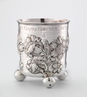  A silver drinking cup titled "Drikkebeger" by Berendt Johansen, featuring elaborate embossed and engraved floral patterns, resting on three spherical feet with a softly gleaming polished finish.