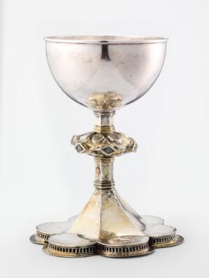  A silver-colored chalice with an ornate stem featuring what appears to be gemstone embellishments, and a richly decorated base, all against a plain white background. Artist name and title remain unknown.