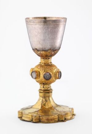  An ornate golden chalice with a slightly translucent silver-grey cup, featuring a decorative stem with blue medallioned knobs, on a neutral background. Artist name and title are unknown.