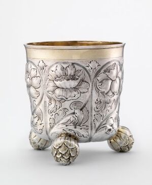  A silver-colored, cylindrical container embellished with embossed floral patterns, with a golden rim on top and resting on three artichoke-style feet.