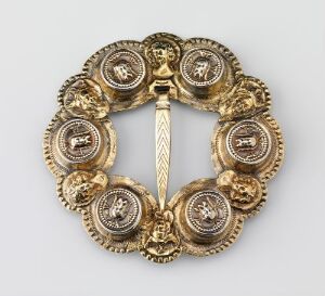  An intricate circular golden brooch or clasp with a symmetrical centerpiece and six detailed roundels showing embossed motifs, crafted in a rich gold that highlights its ornate details and possible heraldic symbolism. Artist name and title are unknown.