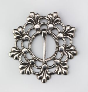  A detailed silver-colored metal ornament, possibly a buckle or brooch, with a symmetrical pattern of stylized petals and spherical motifs arranged in a radial formation around a central hole, featuring polished surfaces and matte recesses for contrast.