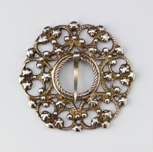  An ornate, round, gold-toned brooch or pendant with an intricate lace-like filigree design, featuring concentric circles at the center and small bead details throughout the pattern.