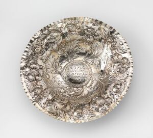  A decorative metal plate featuring a detailed embossed design with a central relief medallion of a face resembling a wind god, surrounded by intricate floral patterns on a raised rim, displaying a silver or pewter-like finish.