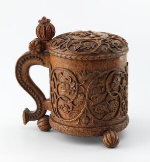  A dark honey-brown lacquered wooden drinking jug, called "Drikkekanne," by an unidentified artist with intricate relief carvings of floral and foliage patterns on its cylindrical body and scroll-shaped handle. The vessel has a decorated lid with a spherical finial.