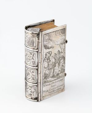  An antique book with a detailed silver cover, featuring narrative engravings and panels, against a plain, off-white background.