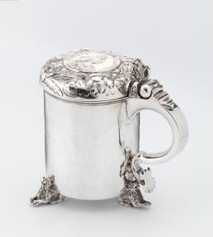  A silver tankard with ornate designs, featuring a cylindrical body, a handle with a scroll-like thumb piece, and four stylized animal paw feet. The lid is elaborately decorated with floral and foliate patterns centered around a circular medallion.