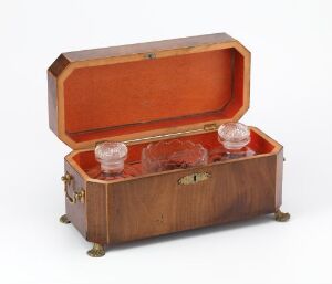  An old small wooden chest with rounded edges and brass detailing, open to reveal two clear faceted glass inkwells in a plush orange-tan lined interior, set against a light gray background.