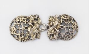  A pair of vintage metal clasps with intricate lace-like cut-out patterns joined together, displaying an aged silver-gray color against a white background.