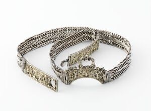  A traditional Norwegian silver bridal belt, "Brudebelte," featuring intricate chain weaving and ornate, partially gilded, cast silver plates with cultural motifs, resting against a light gray background. Artist unidentified.