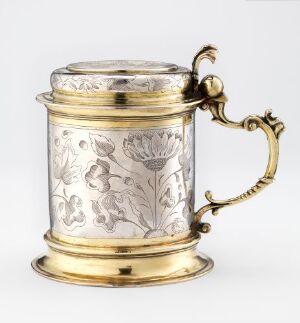  An antique silver tankard with gold accents and a floral etching, featuring a curved gold handle and decorative bands at the top and bottom edges.