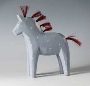  A wooden figure titled "Dalahest" by Katarina Brieditis, depicting a stylized gray horse with white spots, red mane and tail, crafted through wood carving and painting techniques.