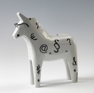  A wooden Dala horse ("Dalahest") by artist Katarina Brieditis, standing upright on its legs, painted white with a variety of black typographic symbols such as "@", "&", "#", "?", and "$" spread across its body.