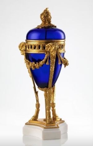  An ornate decorative urn with cobalt blue color and golden embellishments, including a domed lid with a finial, bands with intricate patterns, and three-dimensional gold figures and motifs, displayed on a white base.