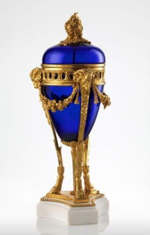  An elegant decorative egg-shaped object with a brilliant royal blue surface accented by ornate gold frameworks, bands, and florals, mounted upon a gold stand against a white background.