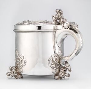  A polished silver tankard with detailed embossed floral decorations on the rim, handle, and base, set against a light grey background. Artist name and title are unknown.