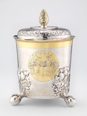  An antique silver mustarding pot with floral engraving and a gold-toned medallion on the front, standing on three scrolled feet with a decorative lid.