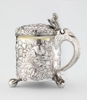  An ornate silver drinking jug titled "Drikkekanne" by Niels Joensen Greger, featuring detailed floral and organic patterns, a vine-like handle, and gold-plated accents standing on three rounded feet.