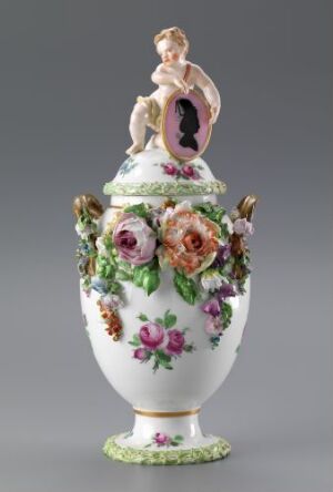  An ornate porcelain urn with a lid, featuring hand-painted flowers in pink, orange, and green, and topped with a molded porcelain cherub in thoughtful pose. The urn has a glossy white finish with a green leafy base.