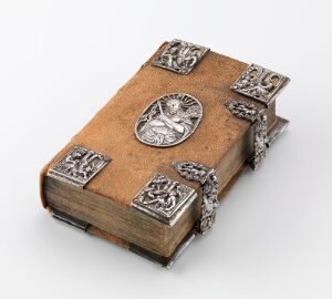  An antique book with a warm beige leather cover, adorned with decorative silver metal elements at the corners and center, resting against a neutral background.